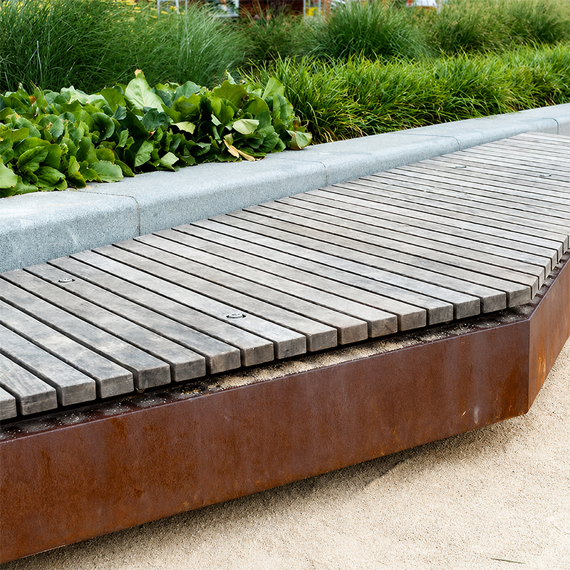 Banc extérieur linéaire avec structure en acier Corten et assise en lames de bois installé près d’un massif végétalisé.