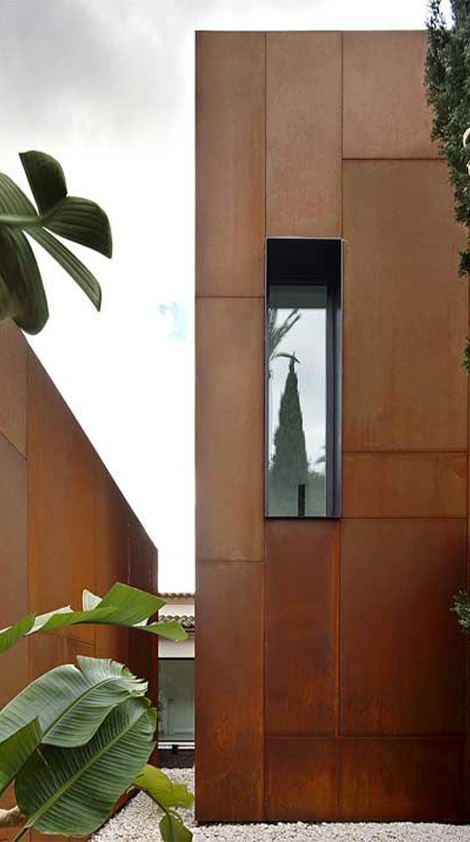 Façade contemporaine habillée de grands panneaux verticaux en acier Corten avec fenêtre étroite et végétation tropicale.