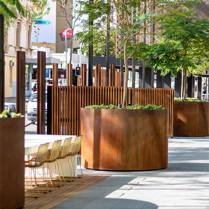 Grandes jardinières cylindriques en acier Corten intégrées à une terrasse urbaine arborée.