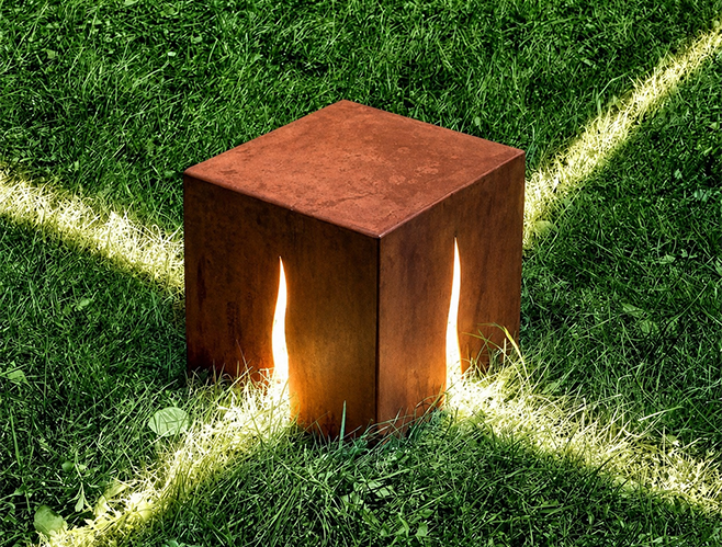 Luminaire cubique en acier Corten installé sur une pelouse avec ouvertures organiques diffusant la lumière.